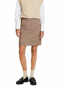 Femme portant un pull sans manches beige, une chemise blanche à manches longues, une jupe à carreaux marron, des chaussettes beiges et des mocassins noirs, debout.