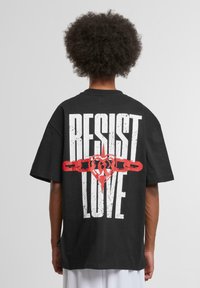 Black oversized t-shirt featuring bold white text "RESIST" and red heart and chain graphic design on the back. Cotton material, short sleeves.