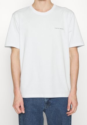 T-shirt blanc à manches courtes avec petit texte "Tiger of Sweden" sur le côté gauche de la poitrine, porté par une personne en jean bleu, vue de face, du cou aux hanches.