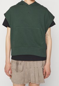 Personne portant un sweat à capuche sans manches vert foncé sur un t-shirt noir et une jupe plissée beige avec une poche latérale et un détail boucle.