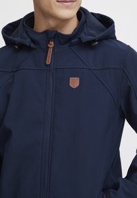 Veste softshell bleu marine avec fermeture éclair à l'avant, capuche réglable, coutures visibles, et un logo en cuir brun sur la poitrine.