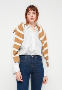 Hvitstripete skjorte med knapper, blå jeans, og en brun og kremstripet strikkegenser lagt over skuldrene.
