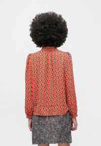 Sarkana blūze ar ģeometriskiem rakstiem, garām piedurknēm un augstu kakla izgriezumu, kombinēta ar teksturētu tumšu svārku.