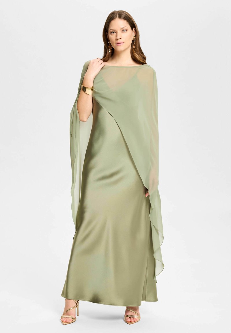 Femme debout portant une robe en satin vert clair avec une cape légère assortie, un bracelet en or, des boucles d'oreilles et des sandales dorées à talons hauts.