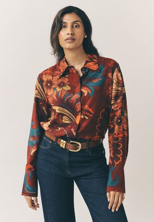 Röd blus med lång ärm och blommigt mönster med blå accenter, kombinerad med mörkblå denimbyxor och ett brunt bälte med guldfärgad spänne.