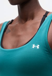 Κοντινό πλάνο ενός ατόμου που φοράει τιρκουάζ αθλητική φανέλα χωρίς μανίκια με ορατό μαύρο ράβδο σουτιέν και λευκό λογότυπο Under Armour στο στήθος.