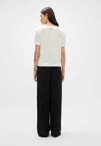 Camiseta de manga corta color crema con un corte relajado, que presenta una textura suave y un pequeño detalle de etiqueta en la parte posterior, combinada con pantalones negros holgados.