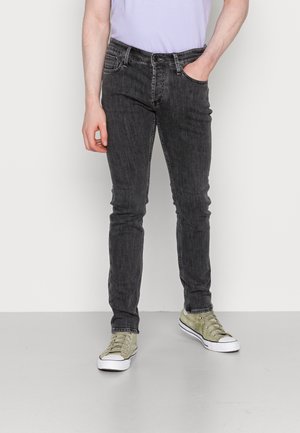 Person trägt ausgewaschene schwarze Slim-Jeans, eine Hand in der Tasche und hellgrüne Leinenschuhe, steht auf grauem Boden.