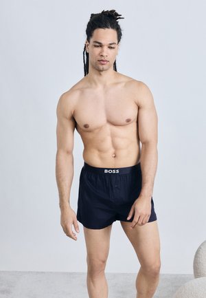 Homme torse nu avec de longues dreadlocks portant un boxer noir BOSS, debout devant un fond gris clair uni.
