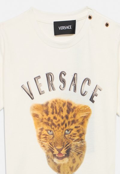 T-shirt crème Versace avec motif visage de léopard, texte Versace au-dessus et deux boutons décoratifs sur l'épaule.