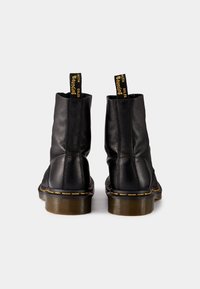 Botas de cuero negro hasta el tobillo con un acabado texturizado, que presentan costuras amarillas en contraste y tiradores en la parte superior para facilitar el calzado.