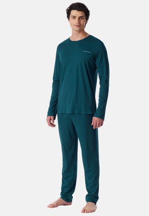 Homme debout pieds nus portant un haut à manches longues couleur sarcelle et un pantalon assorti avec un petit logo texte sur la poitrine sur fond uni.