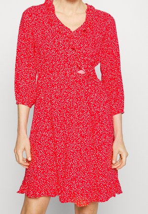 Kvinde iført en rød kjole med hvide abstrakte pletter, trekvartærmer, flæsekant og en let V-udskæring med knapper.