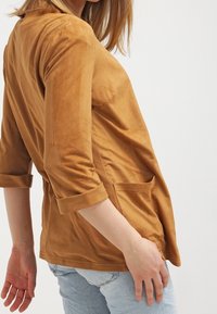 Brun, sammetslen topp med trekvartsärmar, som har en sidficka och uppvikta ärmar, bärs över ljusblå jeans.