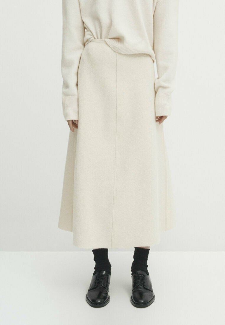 Personne portant une longue jupe crème et un pull, avec des chaussettes noires et des chaussures noires à lacets cirées, debout devant un fond uni.