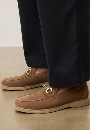 Hnedé semišové mokasíny so strieborným kovovým detailom, nosené s čiernymi nohavicami na béžovej podlahe.