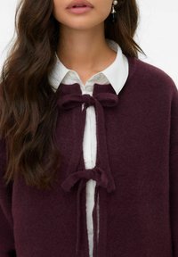 Cardigan bordeaux en tissu doux, orné de deux nœuds liés au niveau du col, superposé à une chemise blanche à col.