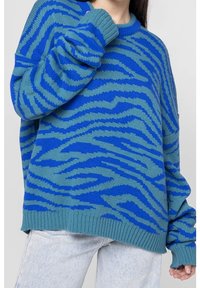 Stickad tröja med ett mönster av blå och teal tigerfläckar, ribbade manschetter och en avslappnad passform, ihop med ljusa denimjeans.