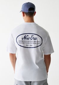 Bijela pamučna majica s okruglim, tamnoplavim logotipom na leđima, koja sadrži natpis "New Era" zajedno s informacijama o njenom porijeklu i godini osnivanja.