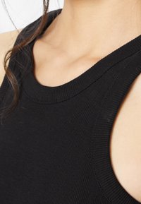 Gros plan d'une personne portant un haut noir sans manches côtelé avec un col rond, montrant la zone du cou et des épaules.