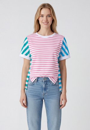 Donna con capelli biondi che indossa una maglietta a righe rosa e bianca con maniche a righe blu e verdi e jeans azzurri, sorridendo in avanti.