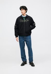 Casaco preto com fecho de correr e logótipo "huf", bordado detalhado nas mangas, combinado com jeans azuis e sapatos pretos. Design simples e de ajuste descontraído.