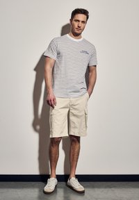 Bílé a námořnické proužkované tričko s kulatým výstřihem, spárované s béžovými cargo šortkami. Tenisky jsou bílé s béžovým detailem na podrážce.