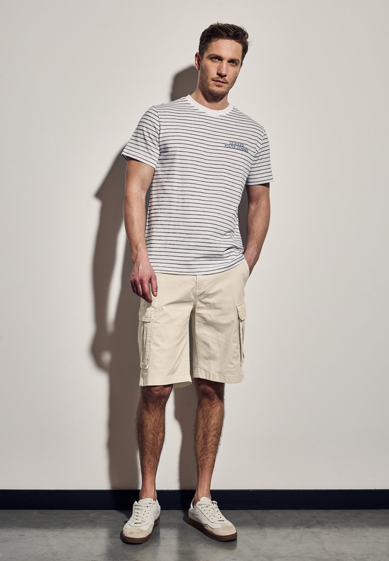 Bílé a námořnické proužkované tričko s kulatým výstřihem, spárované s béžovými cargo šortkami. Tenisky jsou bílé s béžovým detailem na podrážce.