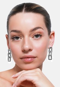 Strieborné náušnice zo skladačiek s prepojenými obdĺžnikovými tvarmi, s lesklým povrchom, nosené modelkou s hladkou pokožkou a prirodzeným make-upom.