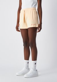 Różowe bawełniane szorty z elastycznym pasem, z logo Champion w pomarańczowym kolorze. Noszone z białymi trampkami i prążkowanymi białymi skarpetkami.