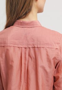 Ljus rosa, lätt skjorta med en slät textur, en kil bak, och en klassisk krage, med sömmar längs kanterna.