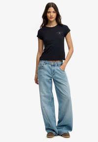 Joven con cabello largo y oscuro, vestida con una camiseta negra corta, jeans anchos de pierna en azul claro y zapatos marrones, de pie con una mano en el bolsillo.