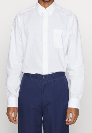 Mann trägt ein weißes, langärmliges Hemd mit Knöpfen und Brusttasche sowie dunkelblaue Hose, steht vor einem schlichten Hintergrund.