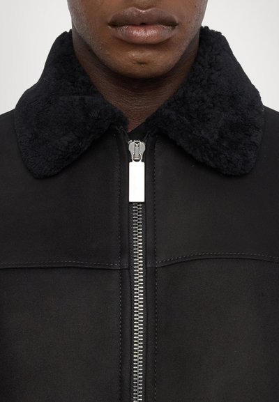 Schwarze Lederjacke mit einem strukturierten, schwarzen Pelzkragen, Frontreißverschluss und sichtbaren Nähdetails. Nahaufnahme des Kragens und des Reißverschlusses.
