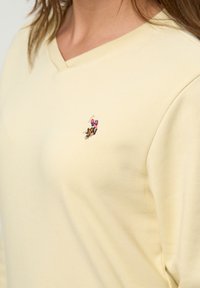 Close-up van een persoon die een lichtgeel V-hals shirt draagt met een klein geborduurd polospelerlogo op de borst.