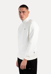 Sudadera blanca con cremallera en el cuello, de textura suave, puños acanalados y un pequeño logo en el pecho. Combinada con pantalones negros.