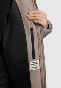 Beige jacka med svart foder, med en ficka med dragkedja och en etikett som säger: "En varm kram gjord av tyg."