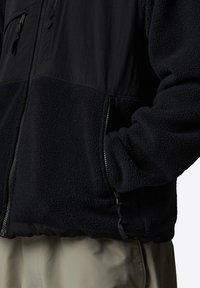Black fleece jacket with a smooth upper panel, textured lower panel, zippered pockets, and elastic cuffs, designed for warmth and comfort.