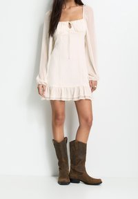 Vestido mini color crema con escote cuadrado, detalle de cordón y mangas largas transparentes, que presenta un dobladillo con volantes. Combinado con botas de cowboy marrones.