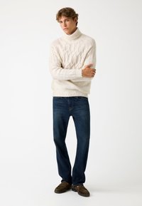 Maglione a collo alto a intreccio crema, vestibilità rilassata, abbinato a jeans blu scuro e mocassini marroni. Design semplice e texturizzato.