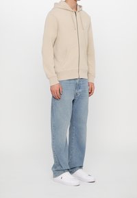Sudadera beige con cremallera y capucha con cordón, dos bolsillos frontales, combinada con jeans de mezclilla azul claro y zapatillas blancas. Ajuste y atuendo casual.