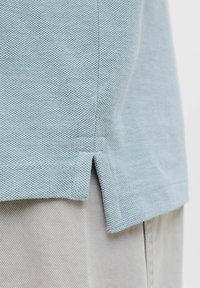 Φως μπλε πουκάμισο από υφασμάτινη ύλη με υφή, με άνοιγμα στο πλάι, πάνω από μπεζ παντελόνια, δείχνοντας λεπτομέρειες του υφάσματος και της ραφής.