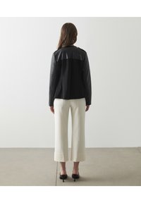 Veste en cuir noir avec une texture lisse et un dos mat contrastant, associée à un pantalon large crème et des escarpins noirs.