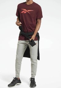 Camiseta marrón con un logo rojo, pantalones de chándal grises y zapatillas negras. Sudadera negra atada a la cintura. Estilo atlético cómodo.