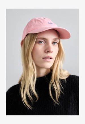 Cappellino da baseball rosa chiaro con ricamo blu navy, visiera curvata e fascia regolabile. Il modello indossa un maglione nero a trama. Lo sfondo è neutro.