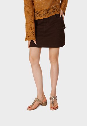 Mujer que lleva una falda marrón con un bolsillo abotonado, un suéter de punto naranja y sandalias beige con adornos decorativos.