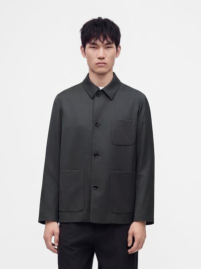 Jeune homme aux cheveux courts noirs porte une veste boutonnée gris foncé avec trois poches avant et un pantalon noir, debout devant un fond blanc.