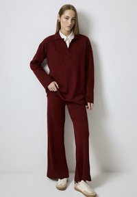 Burgundy knit top with a collar, paired with matching wide-leg pants. Model wears beige sneakers. Smooth texture, relaxed fit.