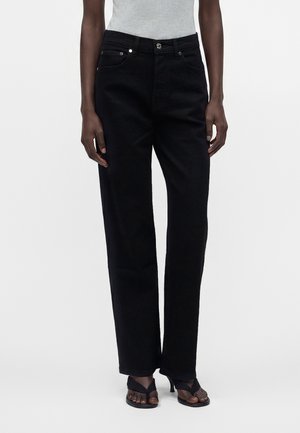 Donna che indossa jeans neri dritti, top grigio senza maniche e sandali neri con tacco e punta aperta, in piedi davanti a uno sfondo semplice.
