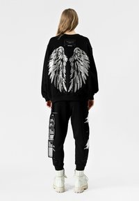 Sudadera negra con diseño de alas plateadas en la parte posterior, combinada con pantalones negros que presentan detalles gráficos y secciones desgastadas. Botas blancas.